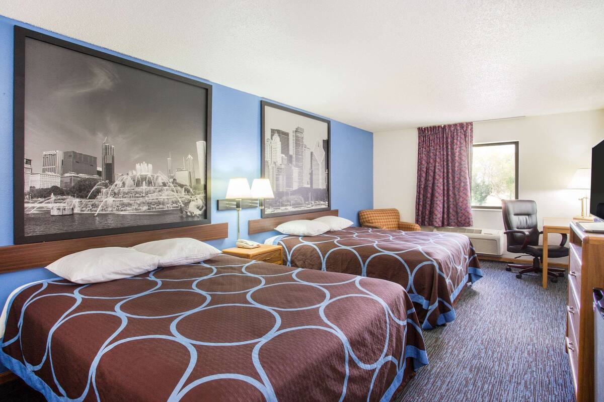 A hotel room with two beds, a desk, a chair, a large window with curtains, and a cityscape view.