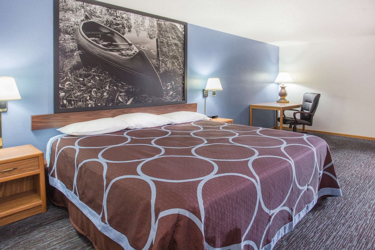 A hotel room with a large bed, a nightstand, a chair, a desk, and a wall-mounted lamp.