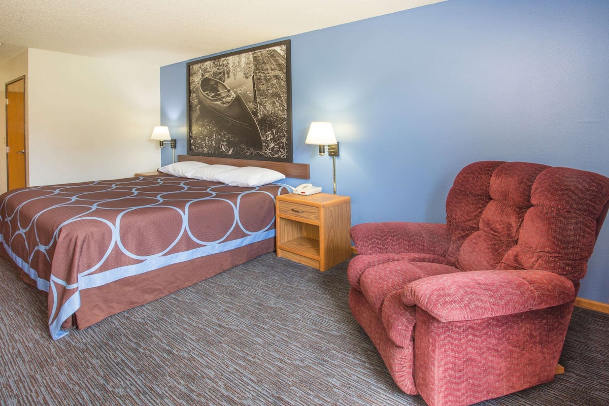 A hotel room with a bed, a red armchair, a nightstand with a lamp, and a wall-mounted artwork.