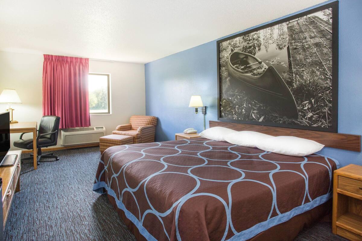 A hotel room with a large bed, a desk with a chair, a small table, a lamp, and a window with curtains.