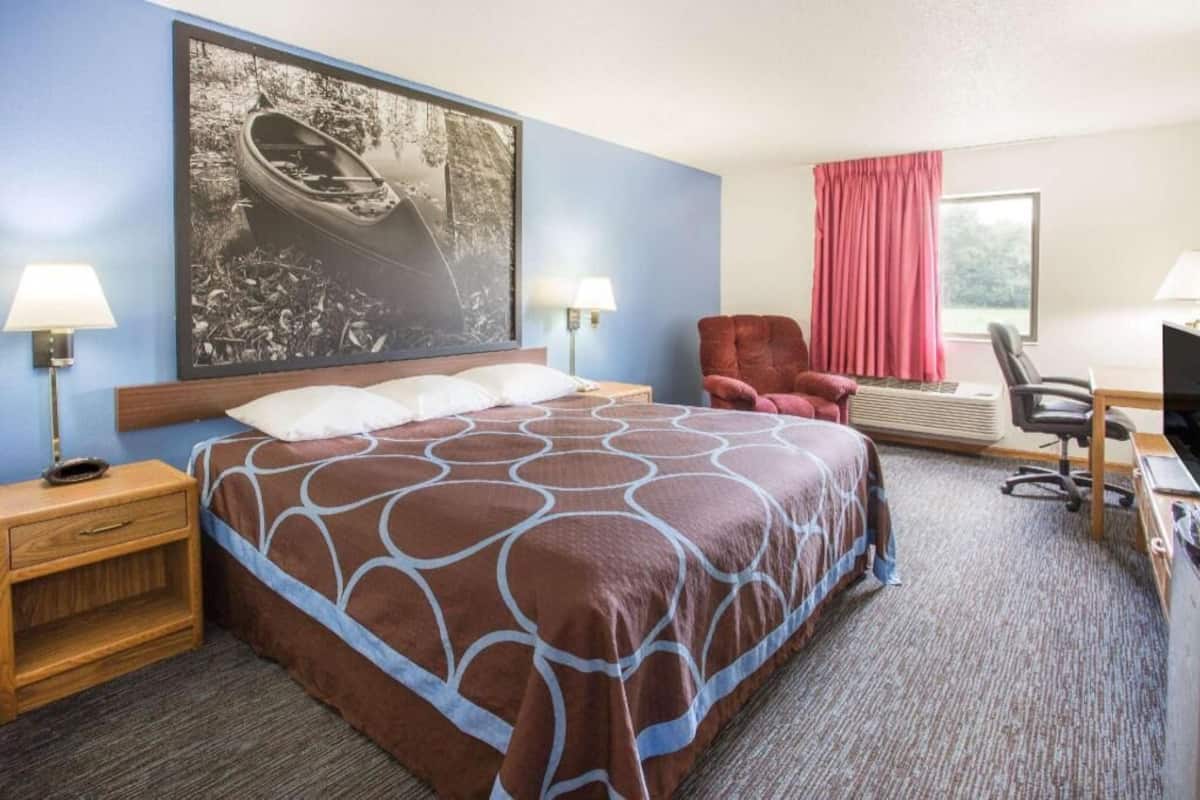 A hotel room with a large bed, two bedside lamps, a desk with a chair, a red armchair, a large framed picture, and a window with pink curtains.