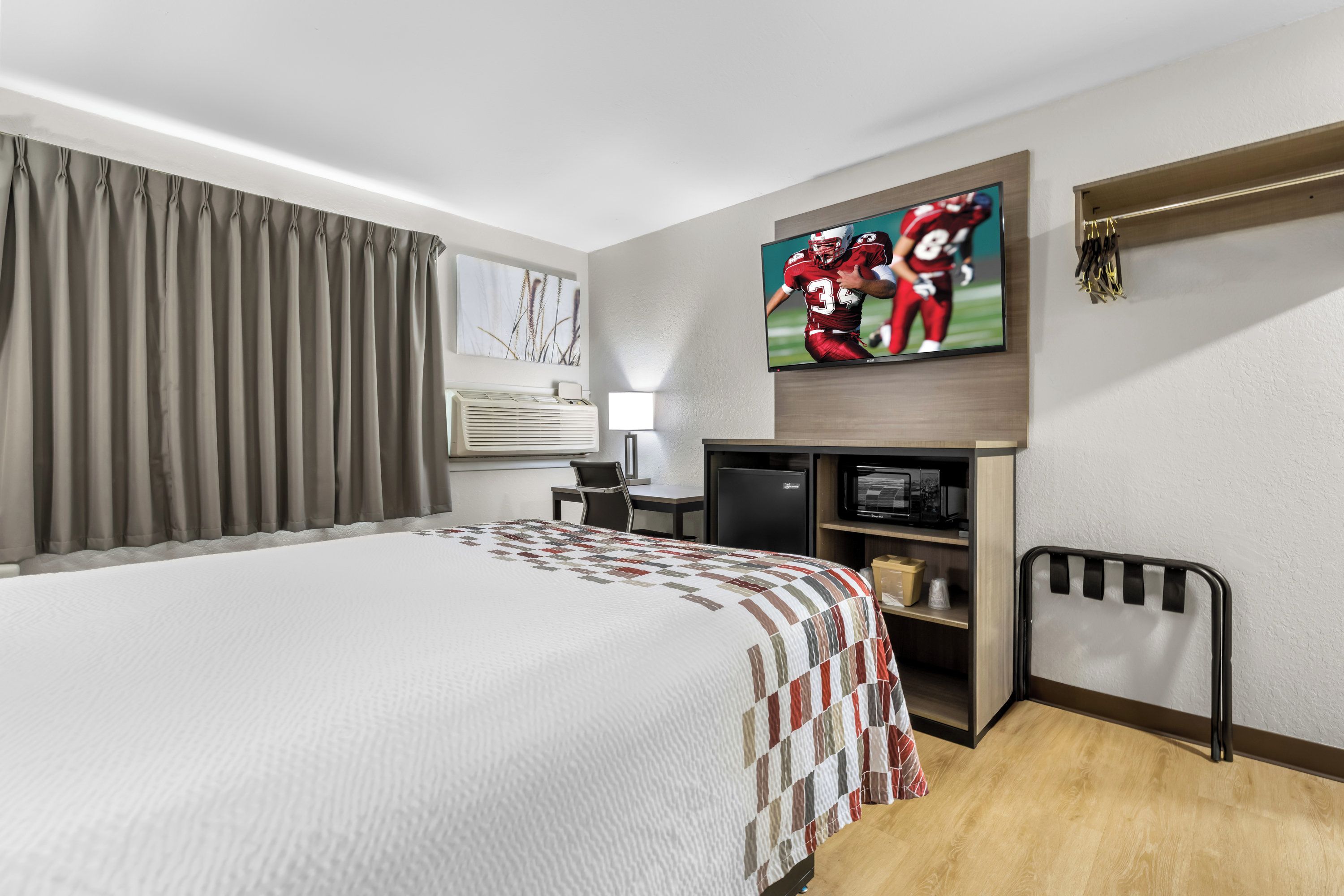 A hotel room with a large bed, a flat-screen TV mounted on the wall, a desk with a chair, a small kitchenette, and a window with curtains.