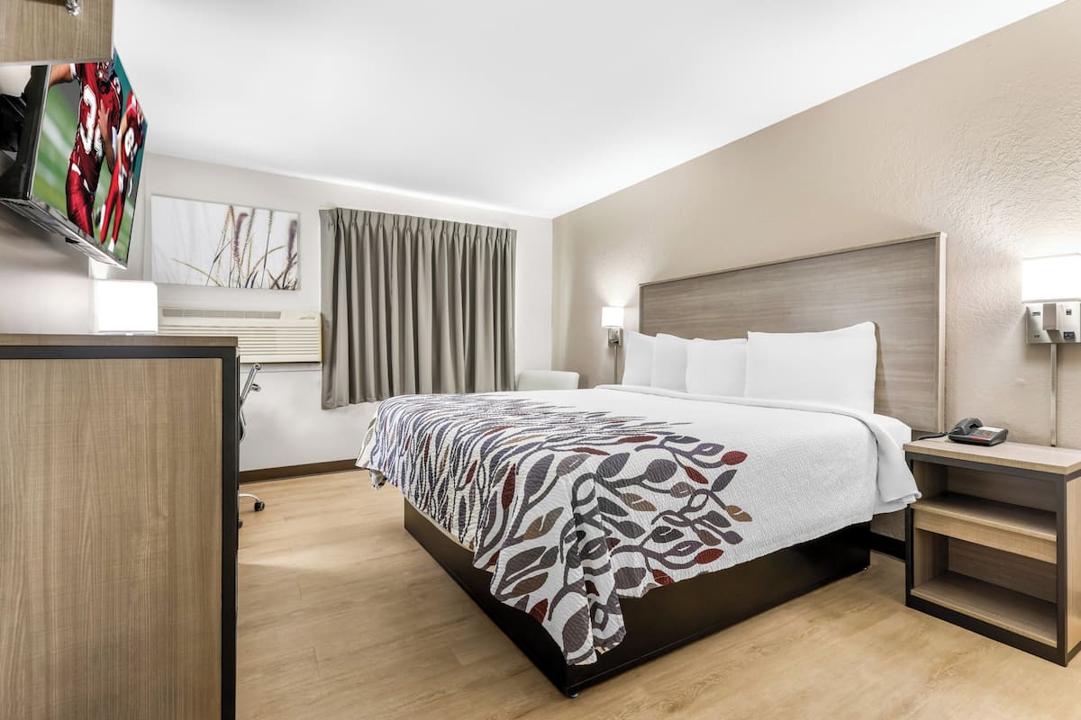 A modern hotel room with a large bed, a bedside table with a phone, a wall-mounted TV, and a window with curtains.