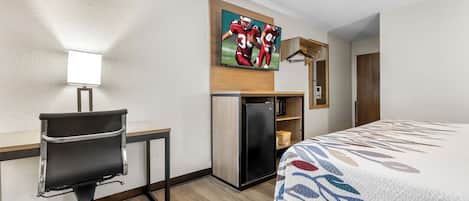 A hotel room with a bed, a desk with a chair, a television mounted on the wall, and a refrigerator.