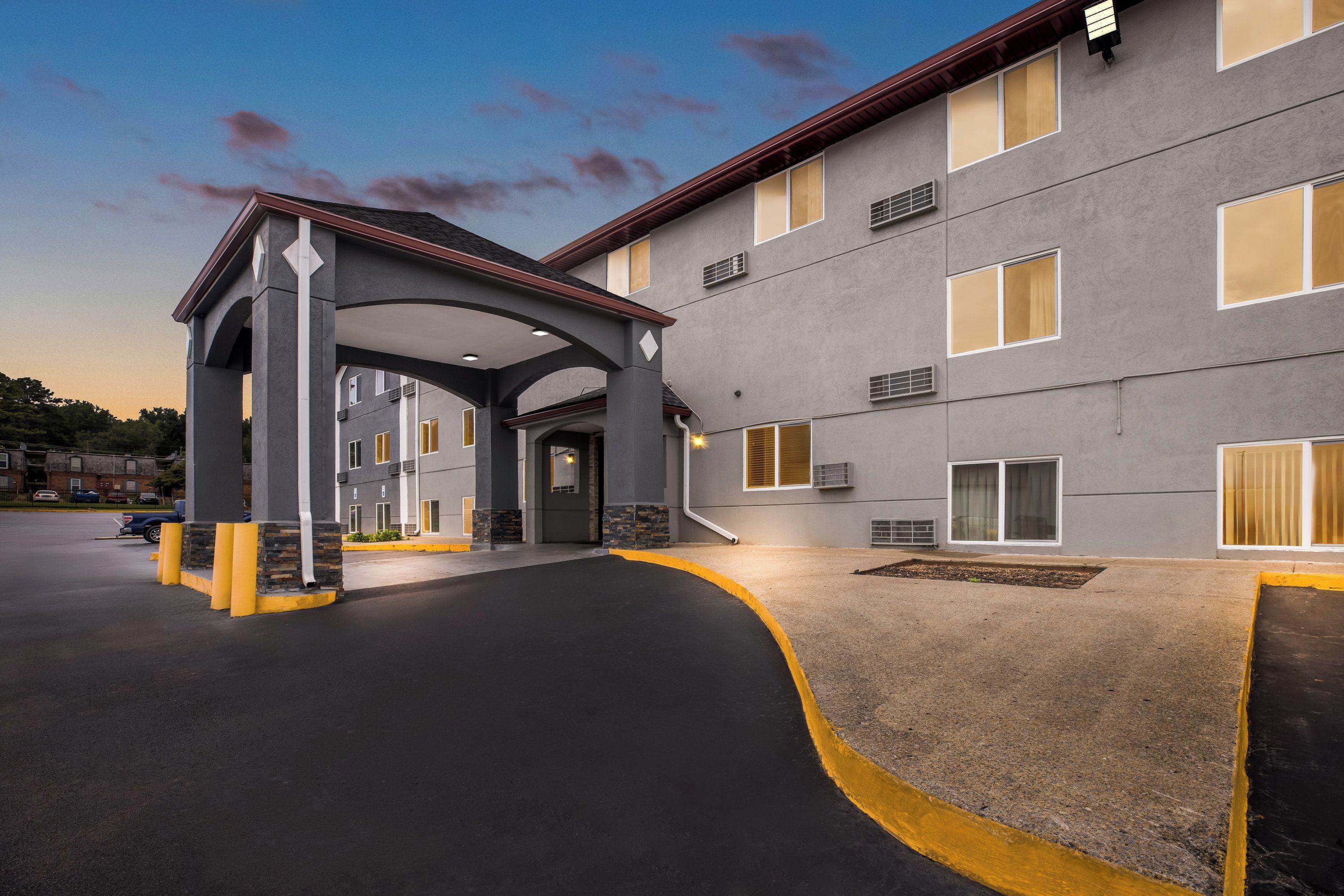 A hotel entrance with a covered walkway, a parking lot, and a building facade with multiple windows and air conditioning units.