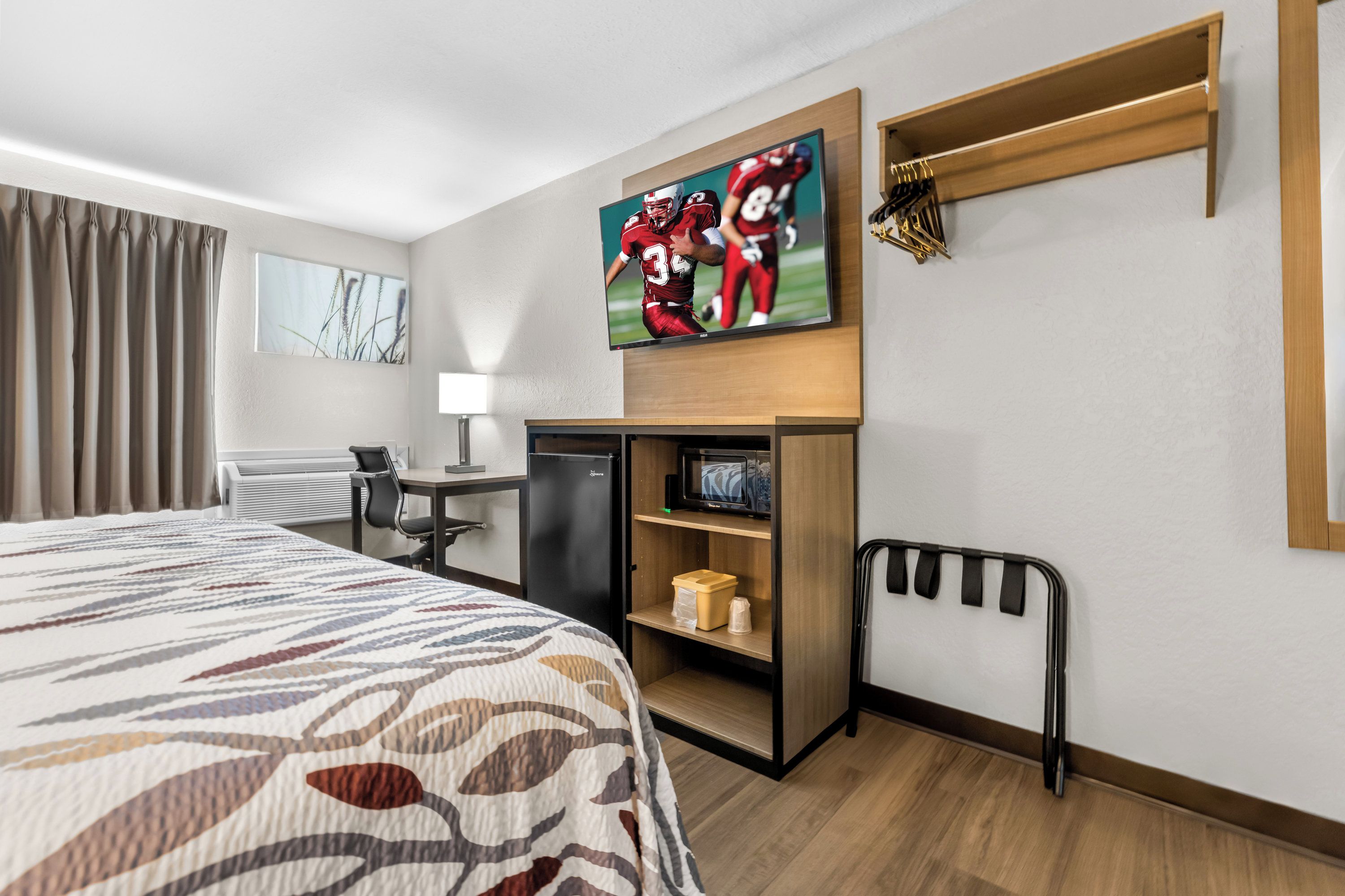 A hotel room with a bed, a desk, a TV mounted on the wall, a microwave, and a small shelf with items.