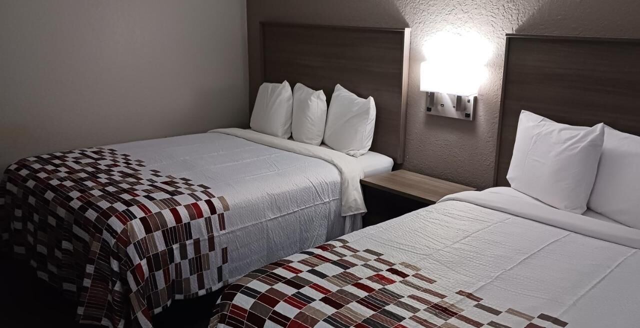 A hotel room with two beds, each with a quilted bedspread, a wooden headboard, and a wall-mounted light fixture.