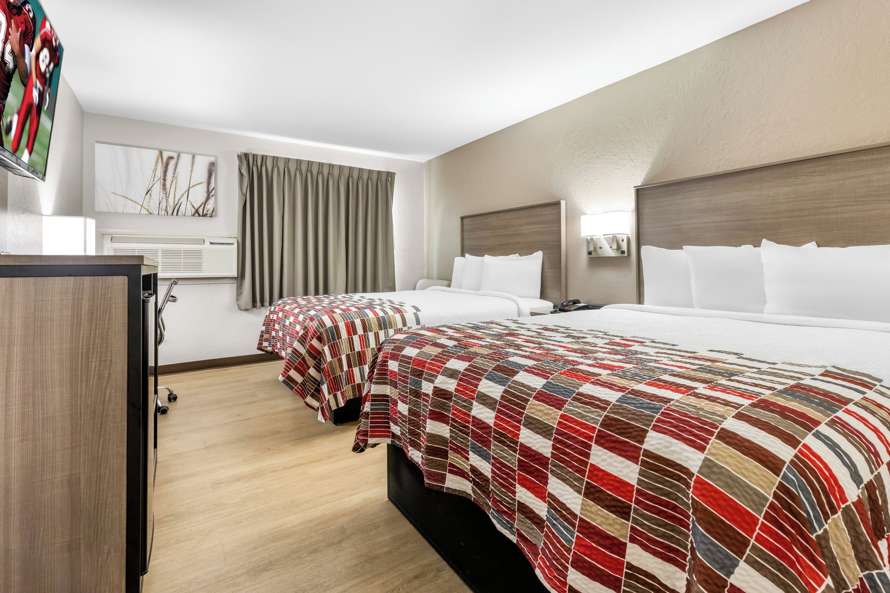 A hotel room with two beds, a TV, a desk, and a window with curtains.