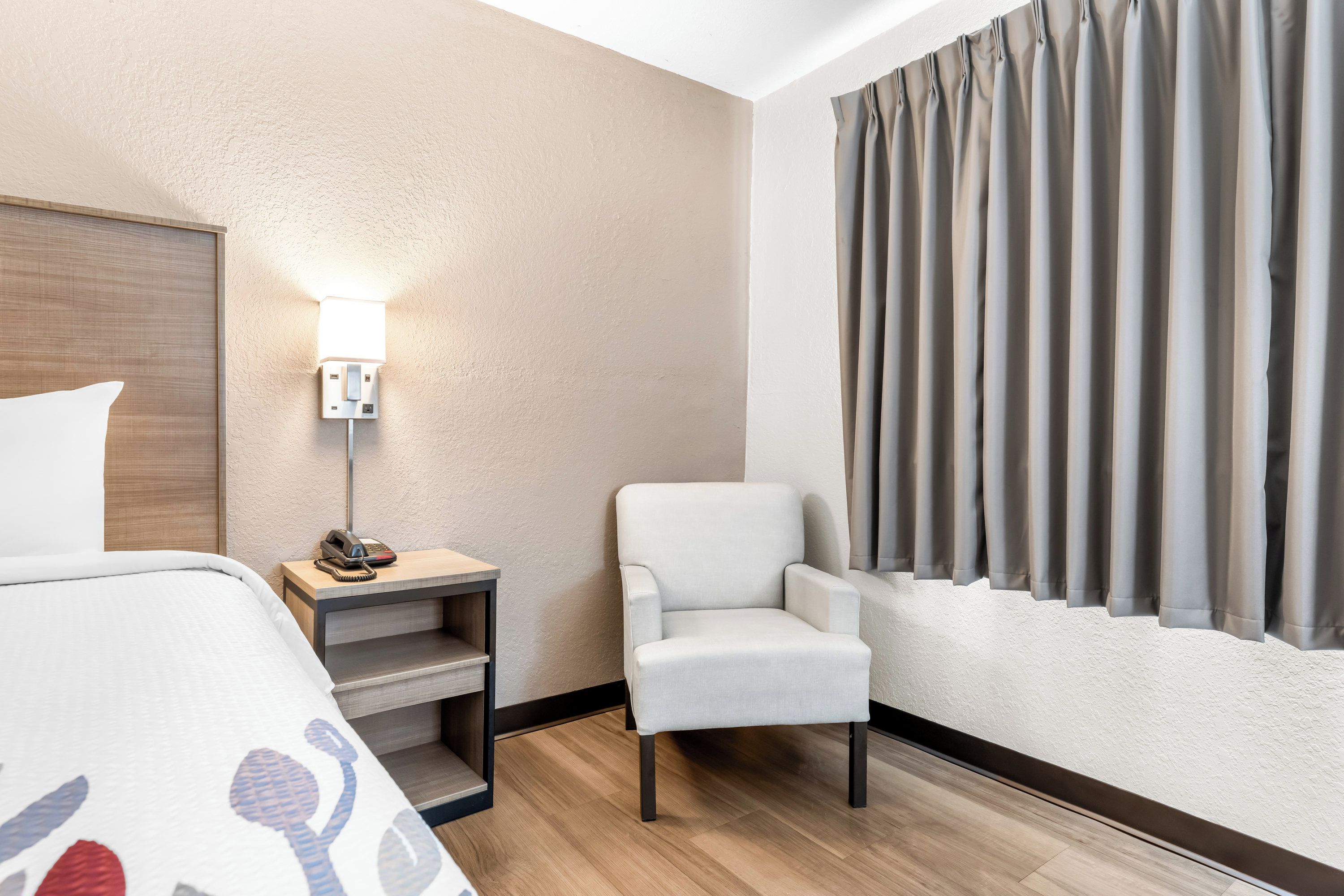 A hotel room with a bed, a chair, a bedside table with a lamp, and a window with curtains.