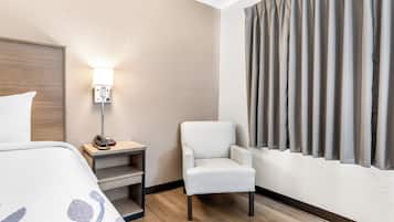 A hotel room with a bed, a chair, a bedside table with a lamp, and a window with curtains.