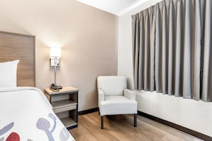 Habitación de hotel con cama, una silla, mesita de noche con lámpara y ventana con cortinas.