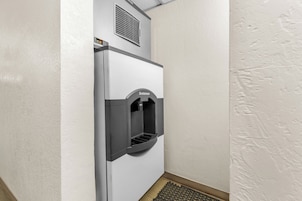 A compact, white, wall-mounted water dispenser with a Scotsman logo, located in a corner of a room with textured walls and a wooden floor.