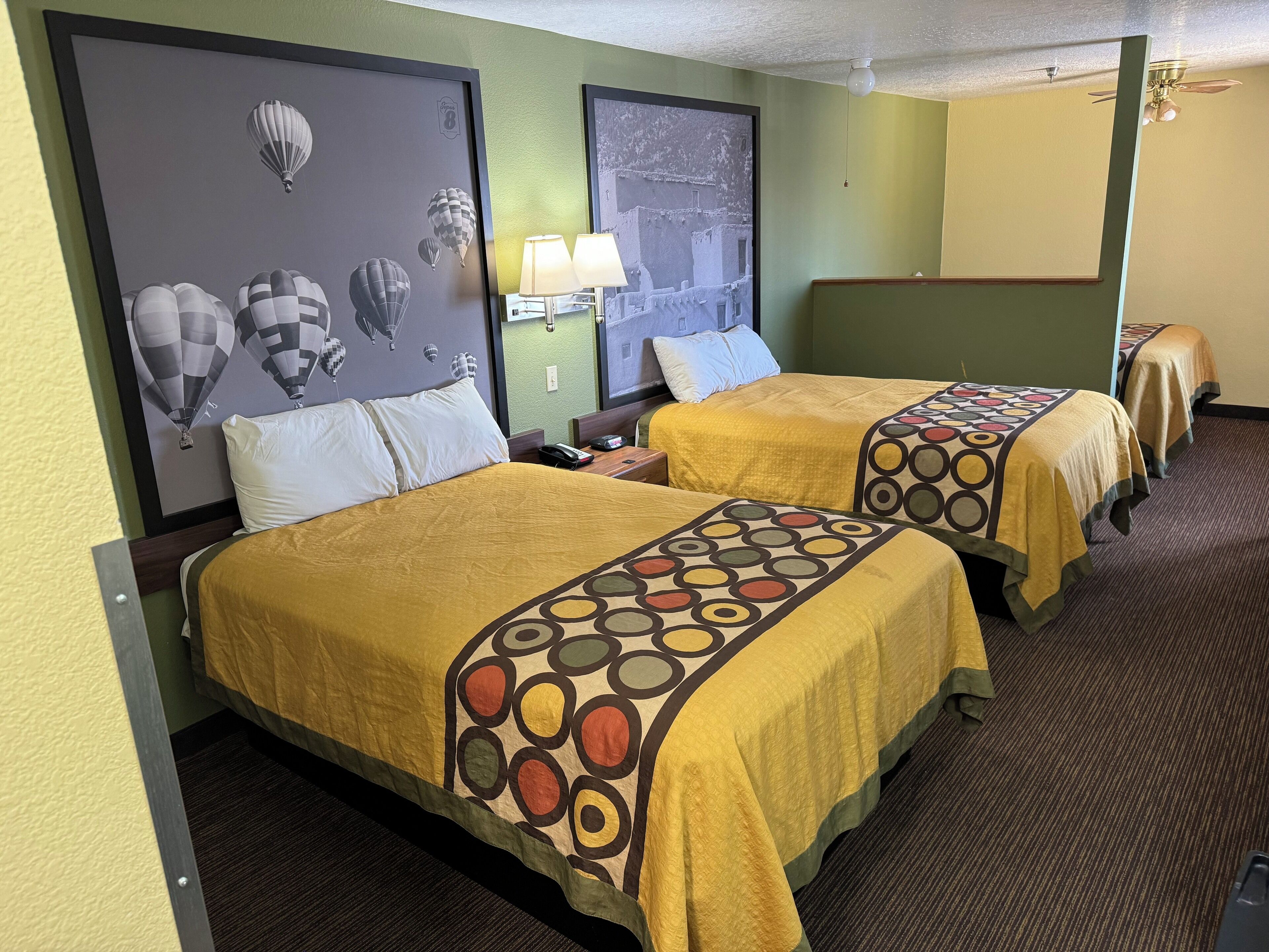 A hotel room with two beds, each with a patterned bedspread, a nightstand with a lamp, and a wall with a framed picture of hot air balloons.
