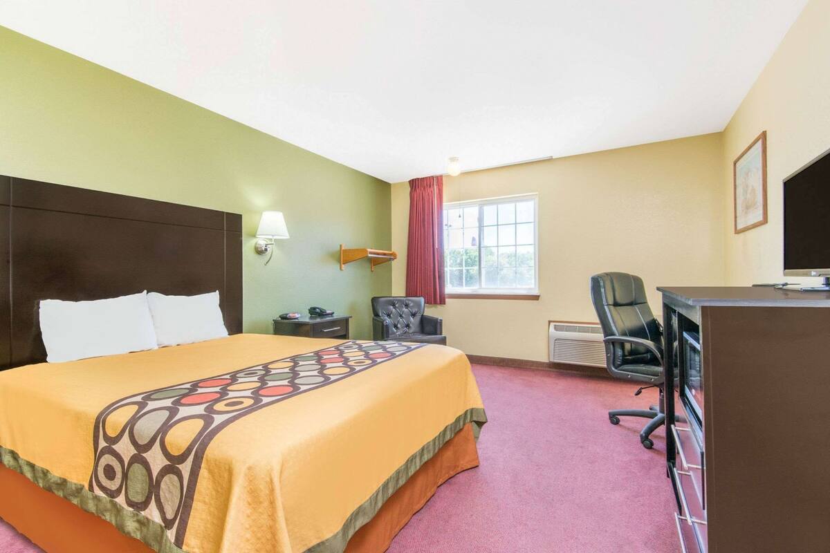 Quarto de hotel com cama, escrivaninha, cadeira, televisão e janela.