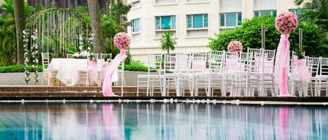 A poolside area with white chairs and tables, pink floral decorations, and a building in the background.