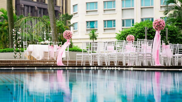 Zona junto a la piscina con sillas y mesas blancas, decoraciones florales rosadas y un edificio al fondo.