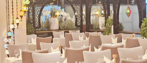 Un restaurante con varias mesas y sillas, elegantes lámparas colgantes y vistas a un jardín a través de un ventanal decorativo.