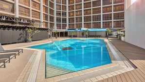 Una piscina en la azotea con suelo de baldosas, sillones de descanso y un edificio grande al fondo.
