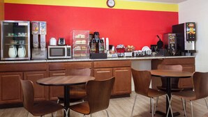 A kitchen with a red and yellow wall, a clock, and various appliances including a refrigerator, microwave, and coffee maker. There are wooden cabinets and two round tables with chairs.