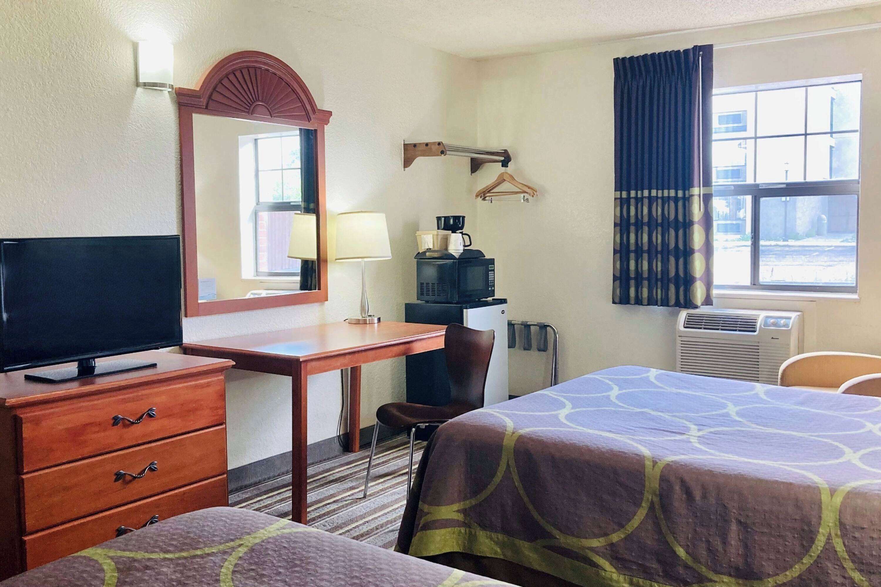 A hotel room with a television, a desk with a lamp, a coffee maker, a bed with a patterned bedspread, and a window with curtains.