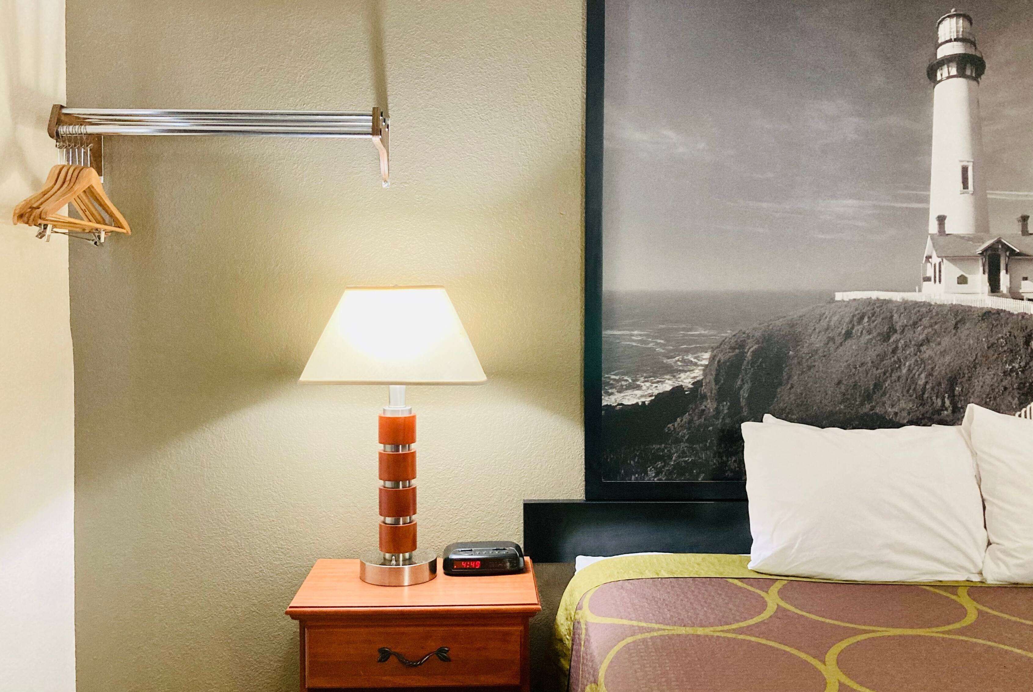 Ein Hotelzimmer mit Bett, Nachttisch mit Lampe, einer Uhr, einem Bilderrahmen mit dem Motiv eines Leuchtturms und einem Kleiderständer mit Kleiderbügeln.