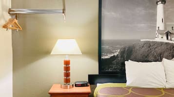 Une chambre d’hôtel comprenant un lit, une table de chevet avec une lampe, une horloge, un tableau représentant un phare, et un porte-manteau avec des cintres.