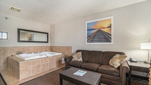 Une chambre d’hôtel avec une grande baignoire, un canapé, une table basse et un tableau représentant un coucher de soleil sur une jetée.