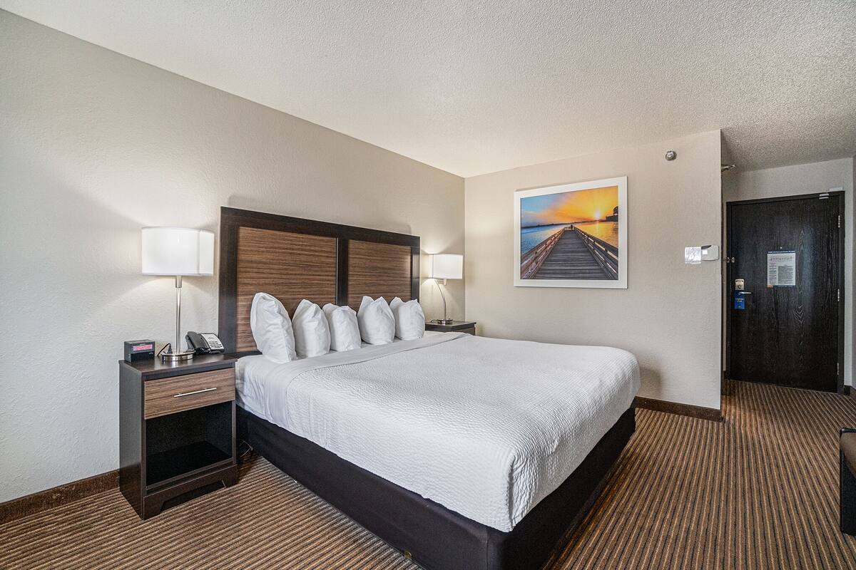 A hotel room with a large bed, two bedside tables with lamps, a nightstand, a painting of a sunset, and a door with a sign.