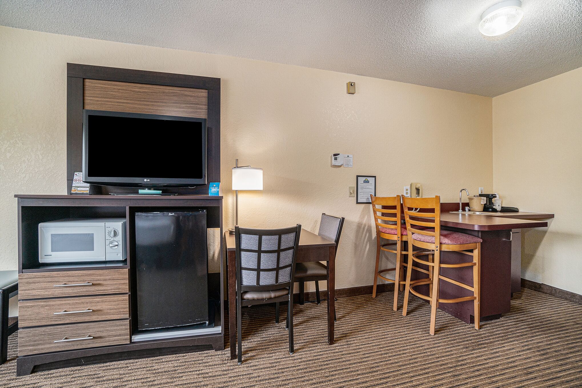 A hotel room with a kitchenette, a flat-screen TV, a microwave, a refrigerator, a dining table with chairs, and a small bar area.