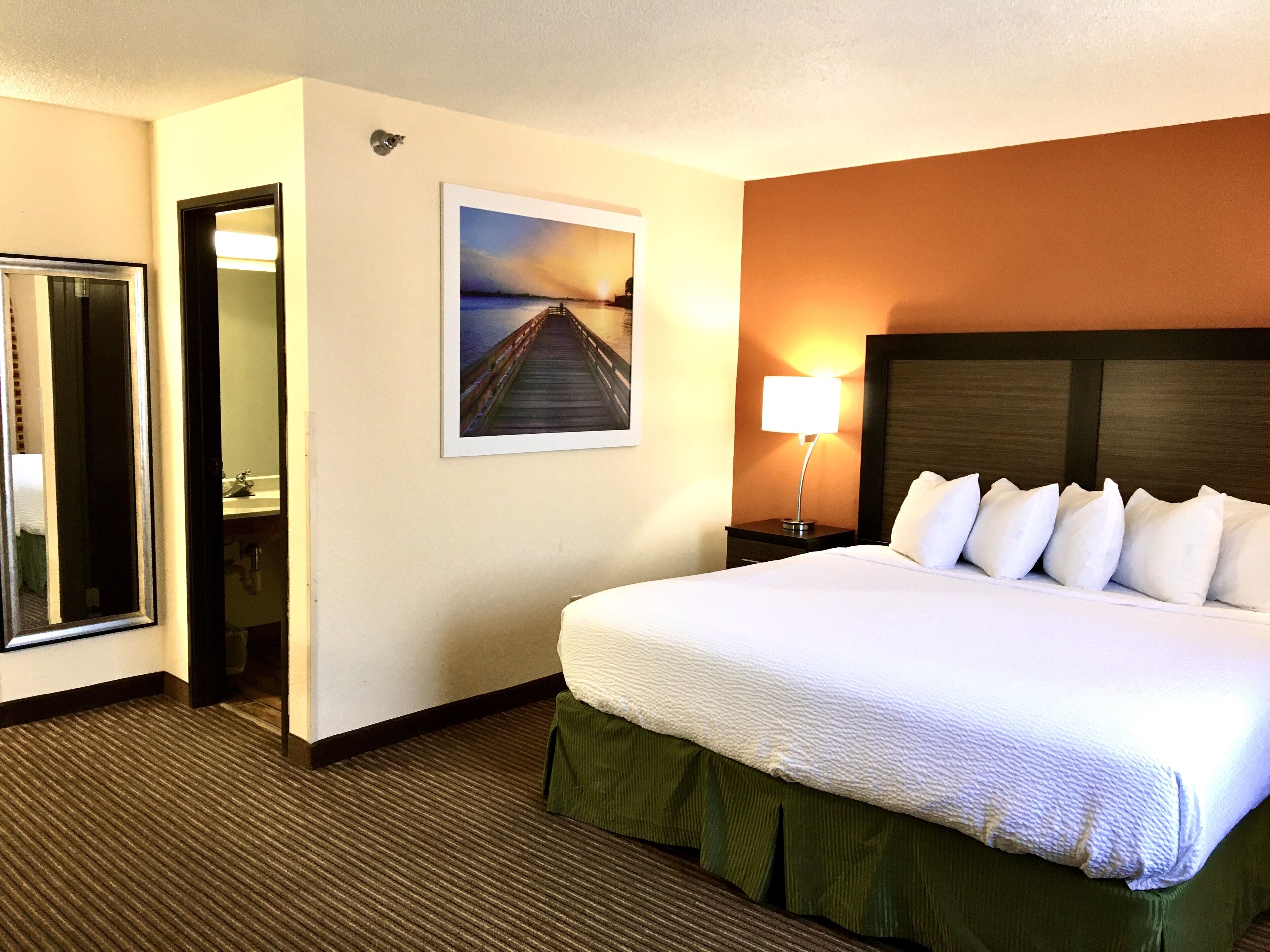 Quarto de hotel com uma cama grande, uma mesa de cabeceira com abajur, um quadro na parede e uma porta para o banheiro.