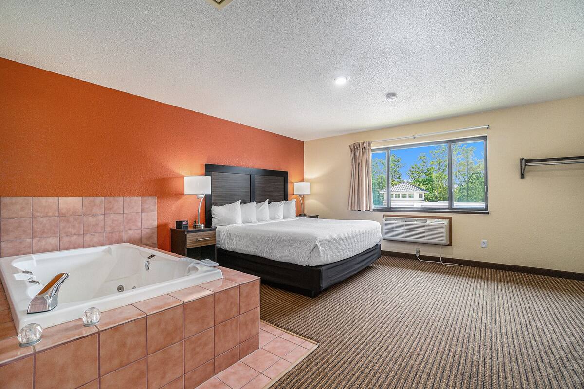A hotel room with a large bed, a separate bathroom with a bathtub, a window with curtains, and a view of trees and a building outside.