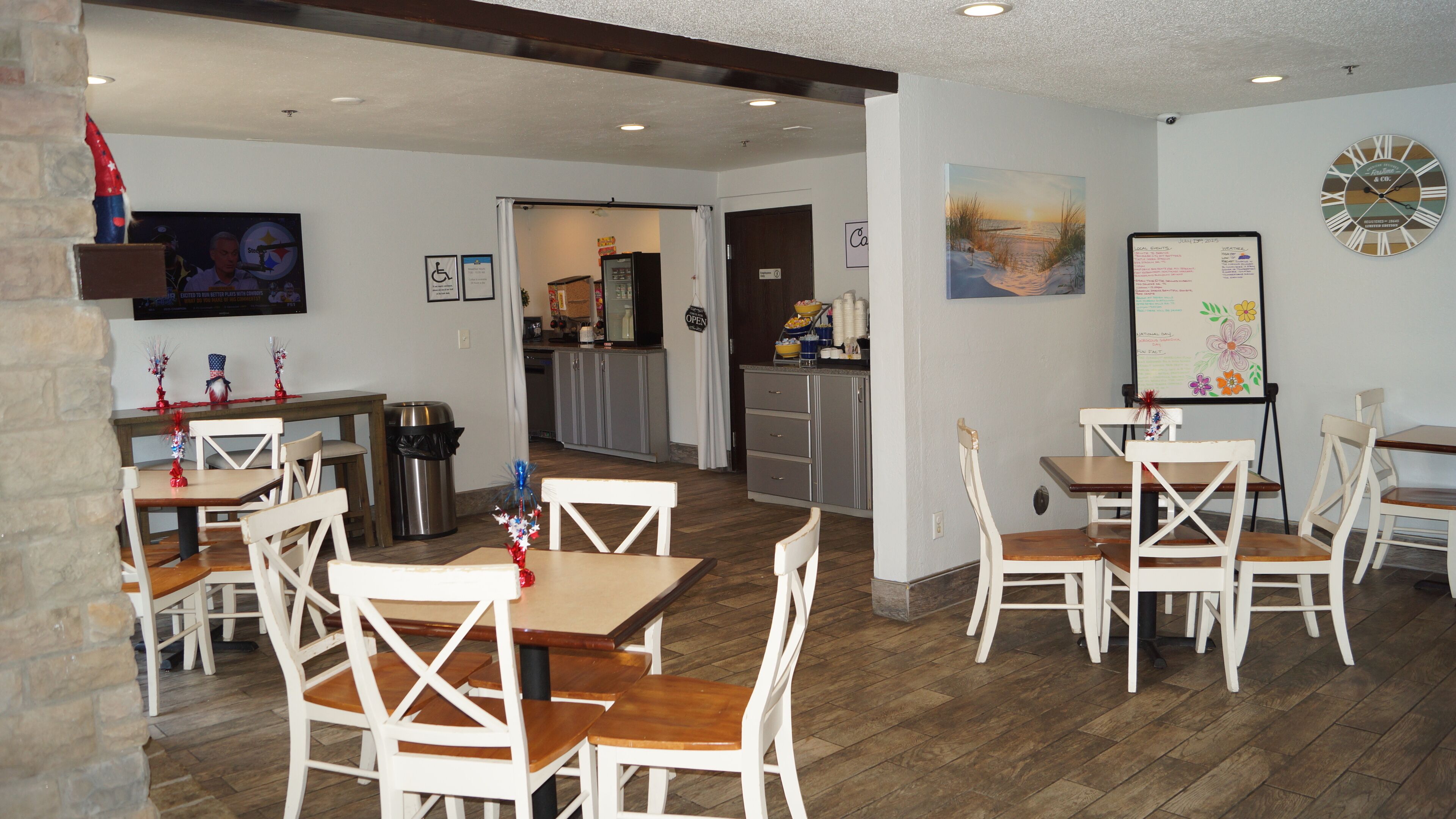 A dining area with wooden tables and white chairs, a kitchen in the background, and a clock on the wall.