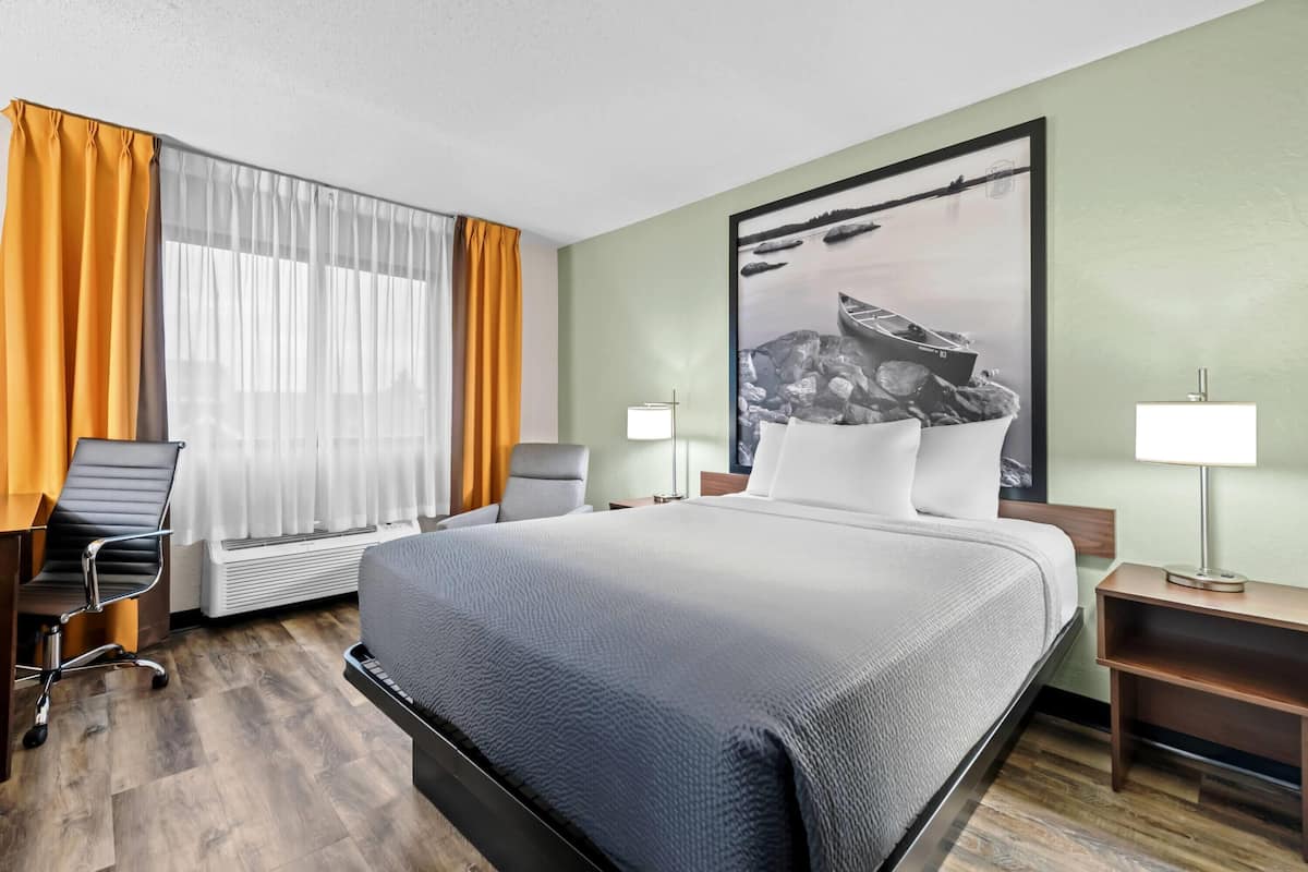 A hotel room with a large bed, a desk with a chair, a standing lamp, and a large framed picture on the wall.