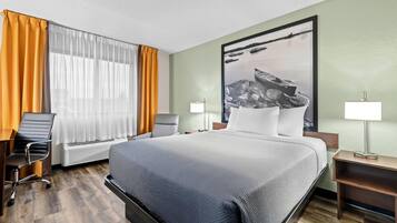 A hotel room with a large bed, a desk with a chair, a standing lamp, and a large framed picture on the wall.