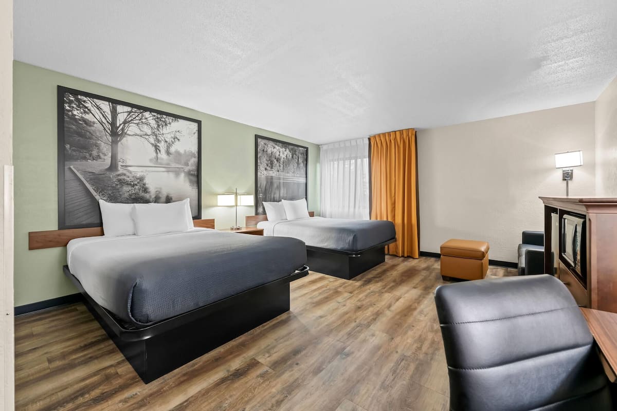 A hotel room with two beds, a desk, a chair, a small table, and a picture of a landscape on the wall.