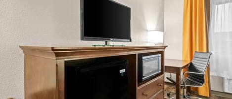 A hotel room with a flat-screen TV mounted on a wooden desk, a mini-fridge, a chair, and a window with curtains.