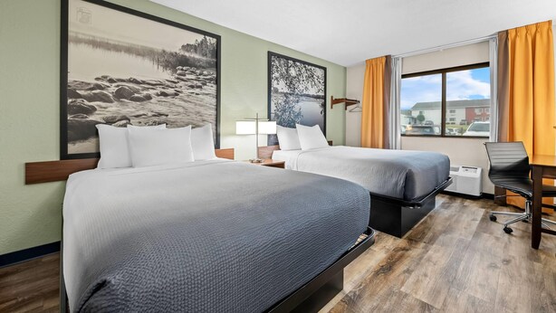 Habitación de hotel con dos camas, un escritorio y una silla. Hay dos cuadros en la pared, una ventana con cortinas y vistas a edificios.