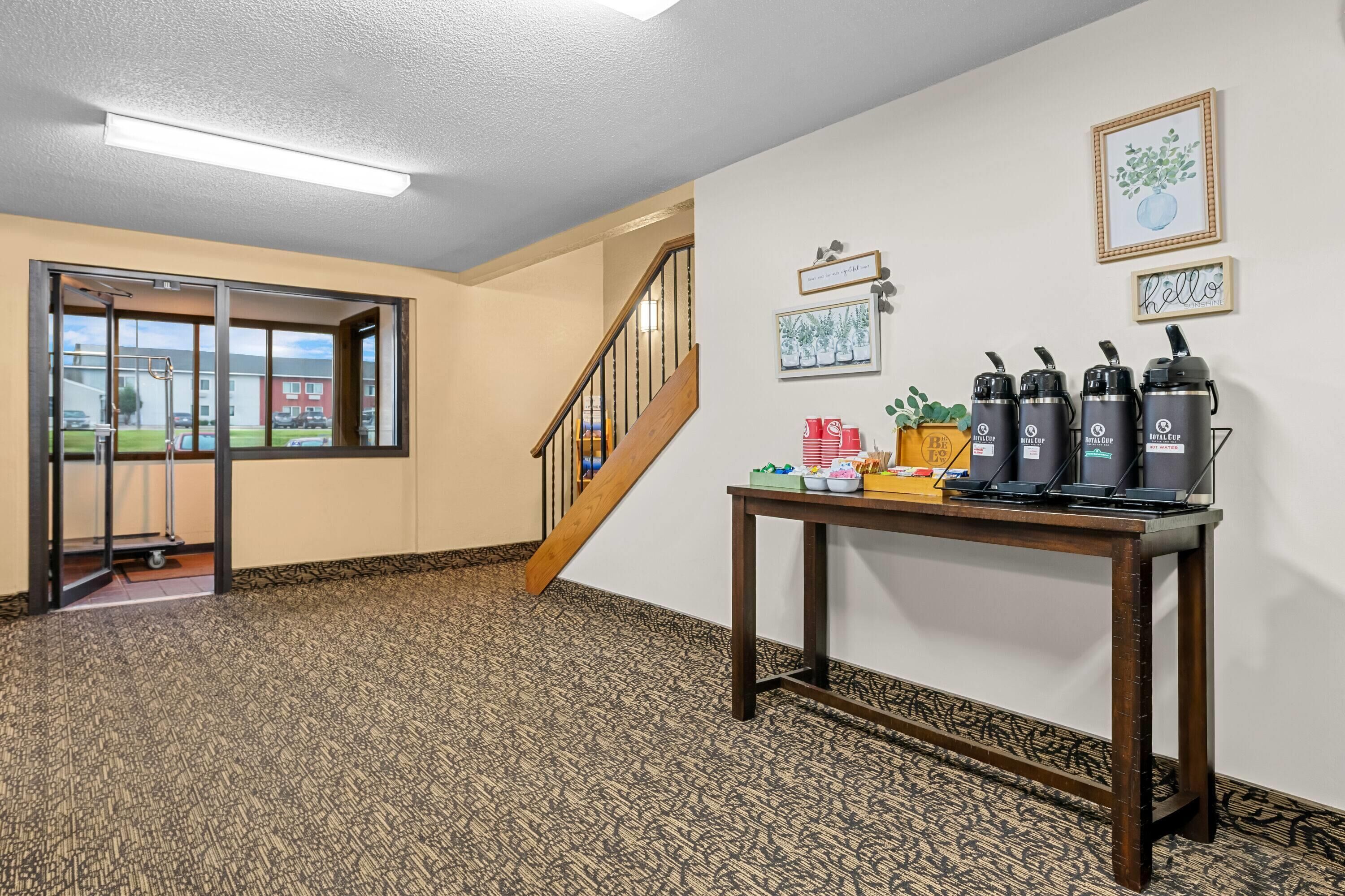 A hotel lobby with a patterned carpet, a wooden staircase, a table with thermos bottles, and framed artwork on the walls.