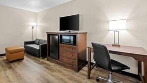 Habitación de hotel con un escritorio de madera, una silla de oficina negra, un sofá, un televisor, un microondas y un pequeño banquito naranja.