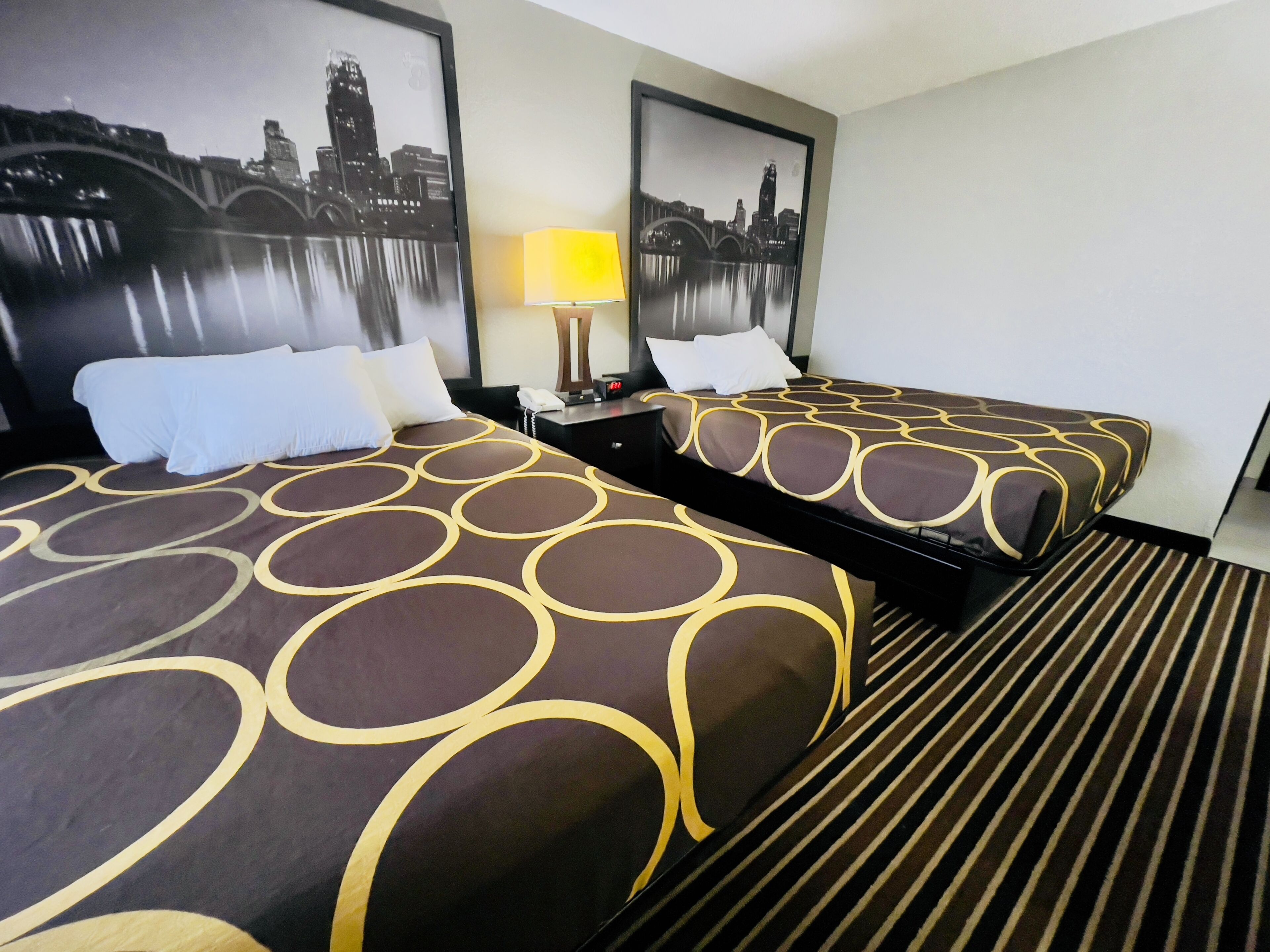 Habitación de hotel con dos camas, cada una con una colcha estampada, una mesita de noche con lámpara y cuadros con vistas urbanas en la pared.