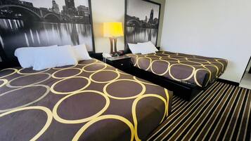 Habitación de hotel con dos camas, cada una con una colcha estampada, una mesita de noche con lámpara y cuadros con vistas urbanas en la pared.