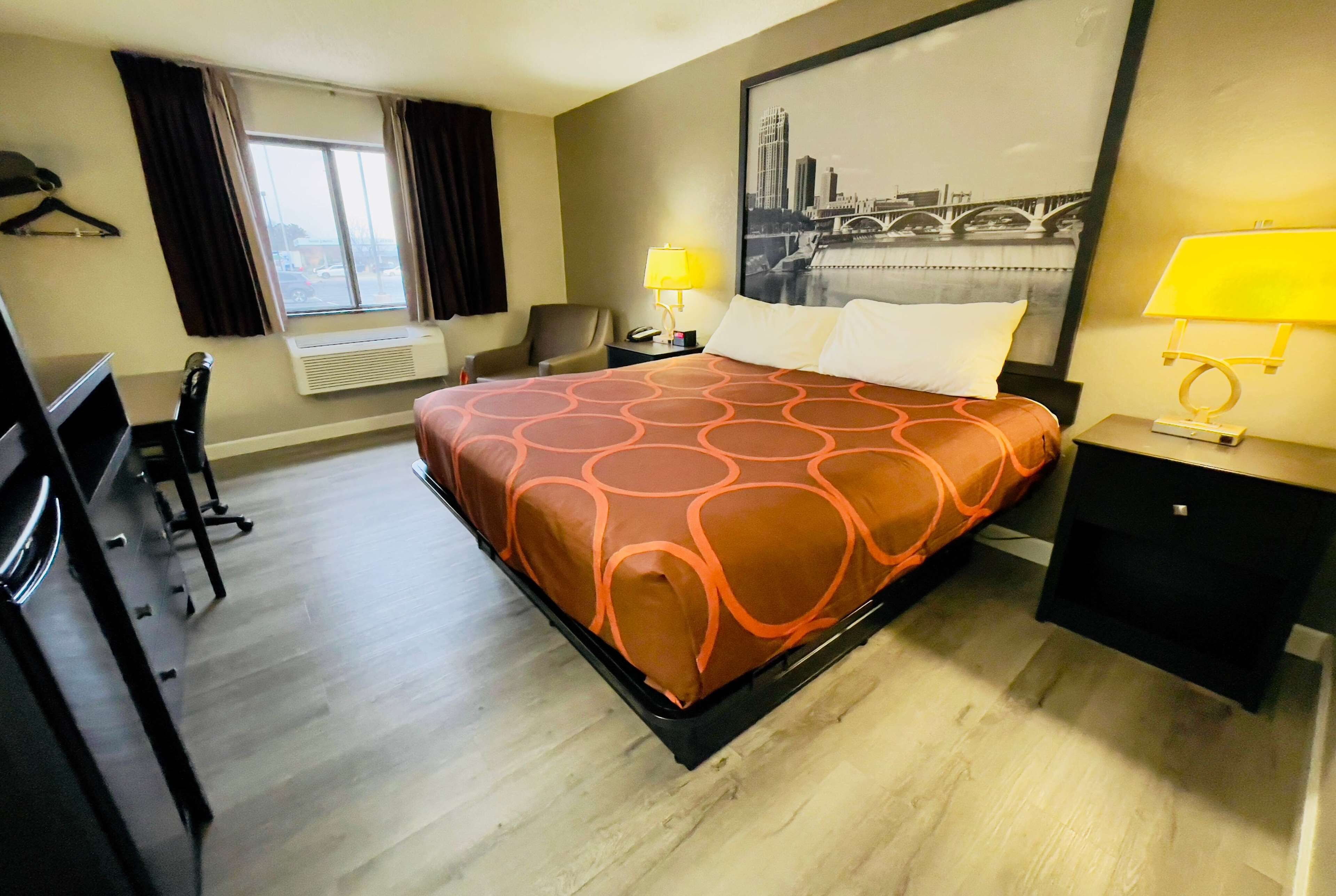 Habitación de hotel con cama, dos mesitas de noche con lámparas, una silla, escritorio con silla, ventana con cortinas y un cuadro grande de un paisaje urbano en la pared.