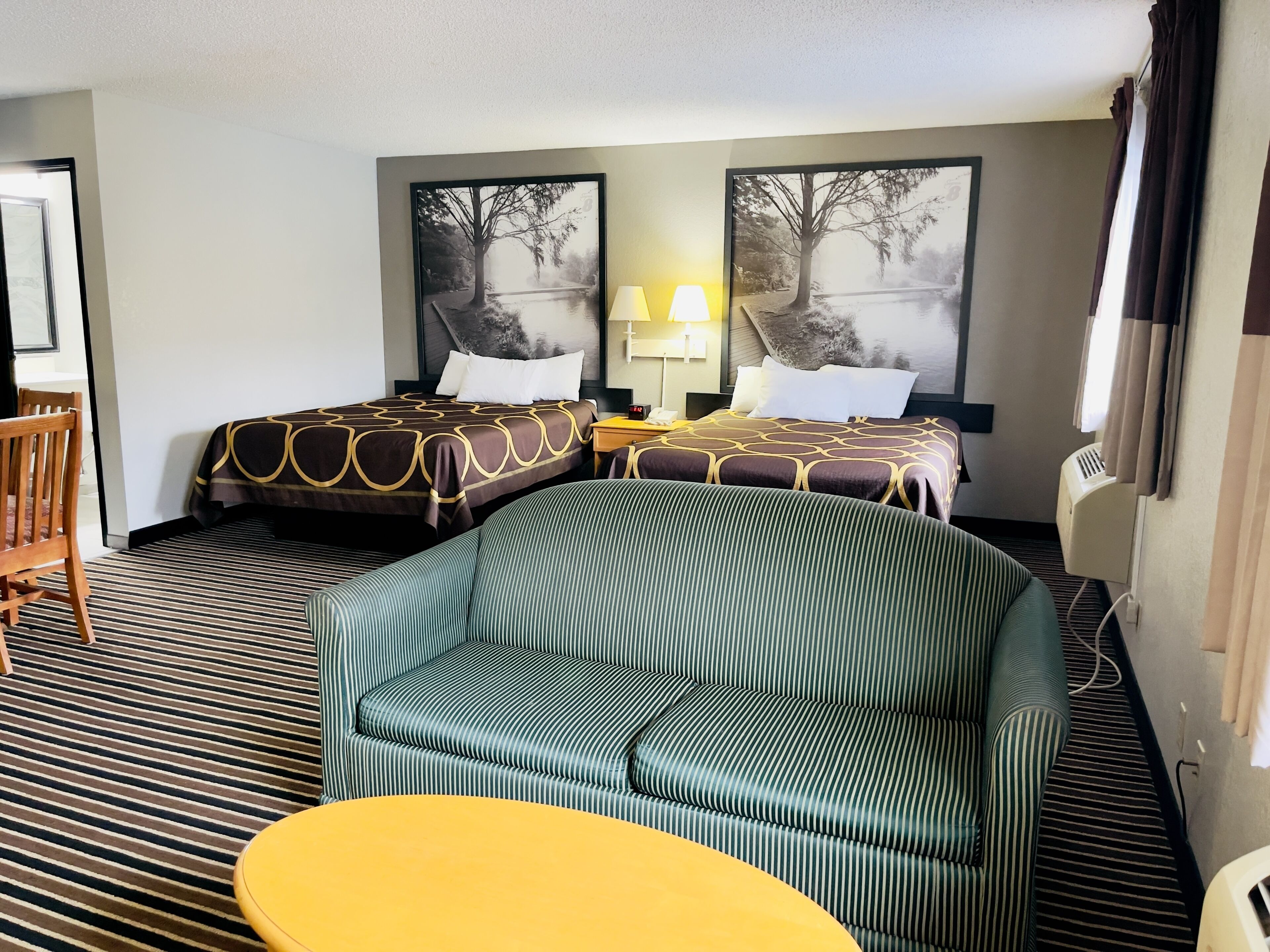 A hotel room with two beds, a sofa, a small table, and a nightstand with a lamp.