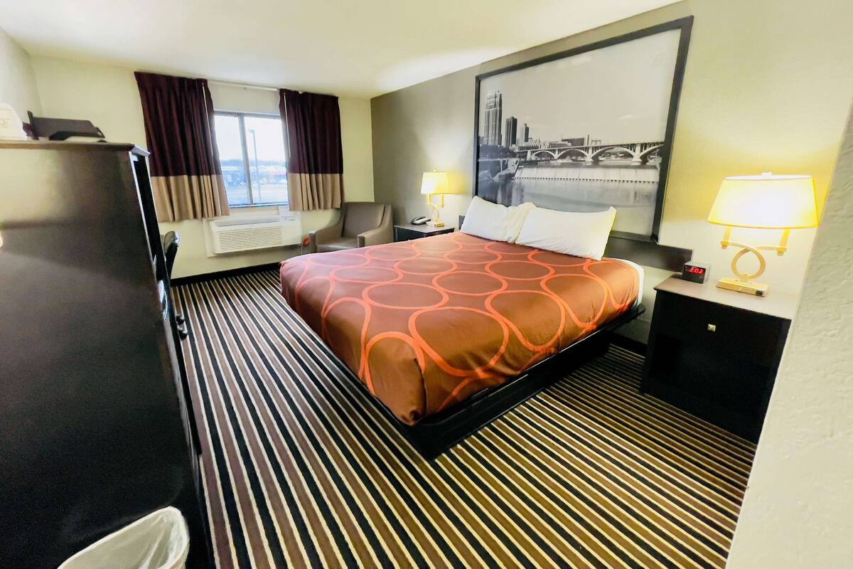 A hotel room with a bed, a desk, a chair, a refrigerator, a window with curtains, and a cityscape painting on the wall.