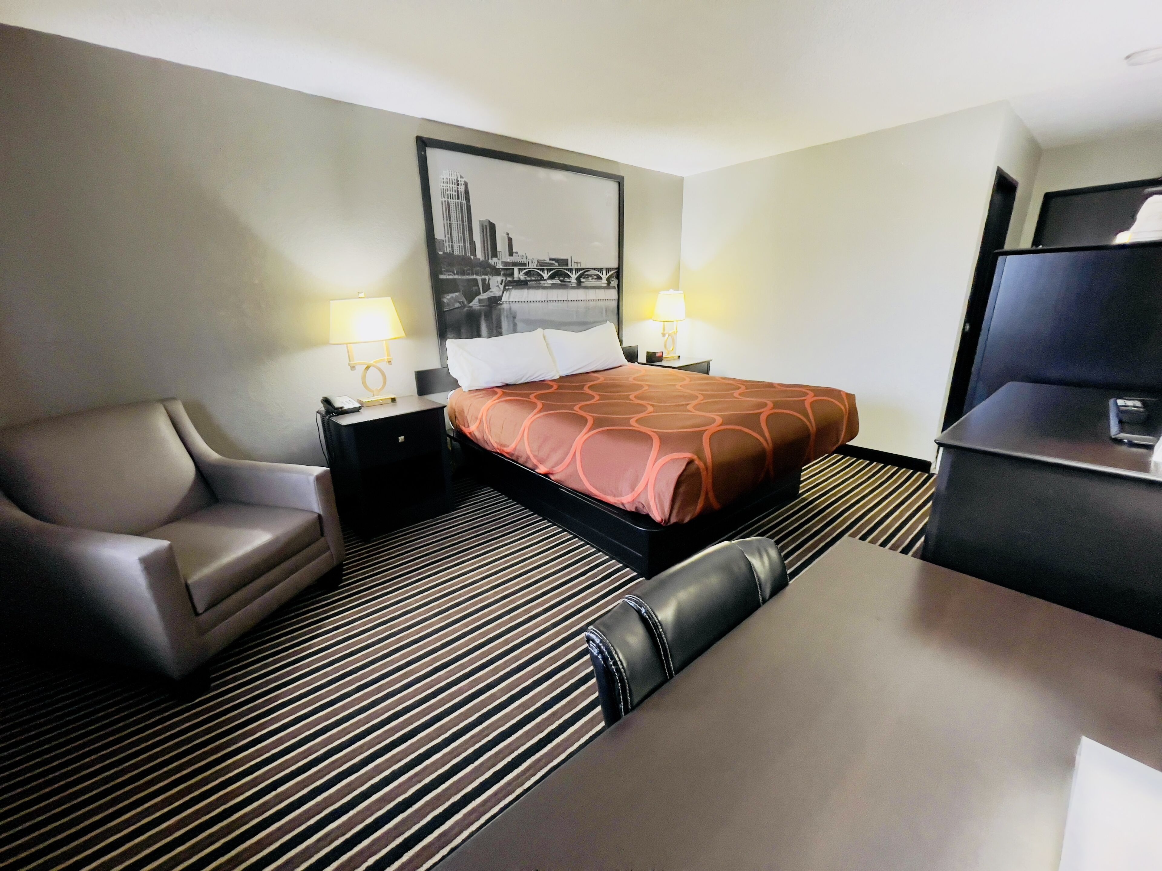 Habitación de hotel con cama, escritorio, silla, dos mesitas de noche con lámparas y un cuadro grande en la pared.