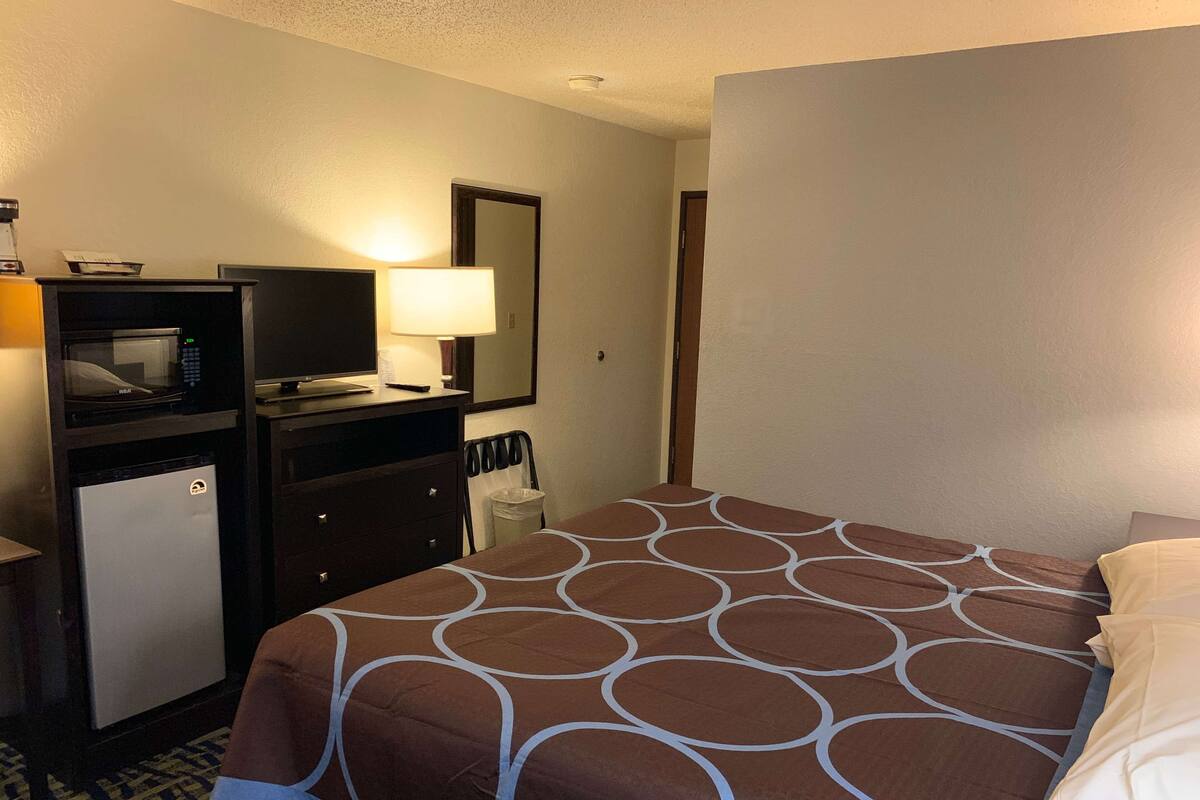 A hotel room with a bed, a desk with a computer and a lamp, a small fridge, and a mirror.