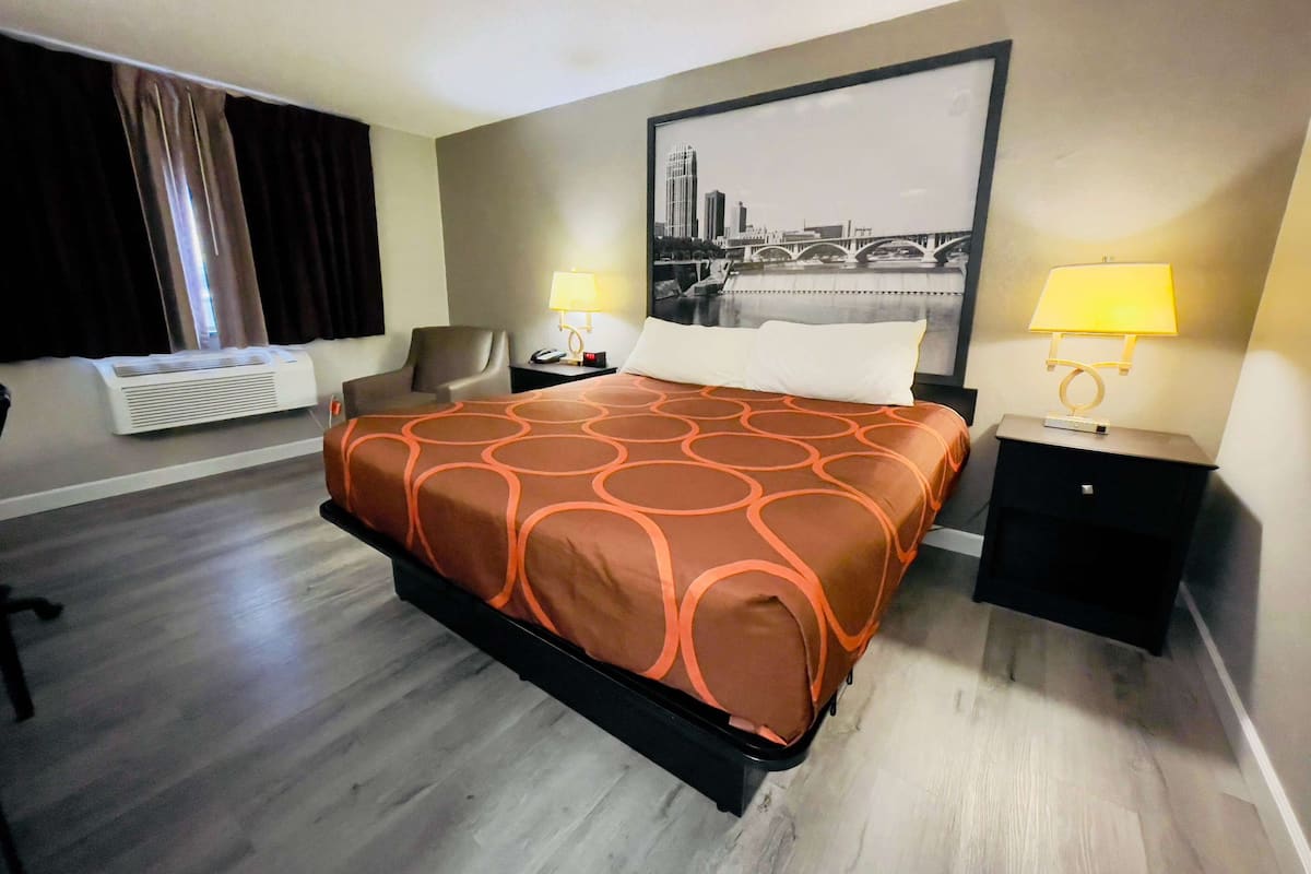 A hotel room with a large bed, two bedside tables with lamps, a chair, a window with curtains, an air conditioning unit, and a cityscape painting on the wall.