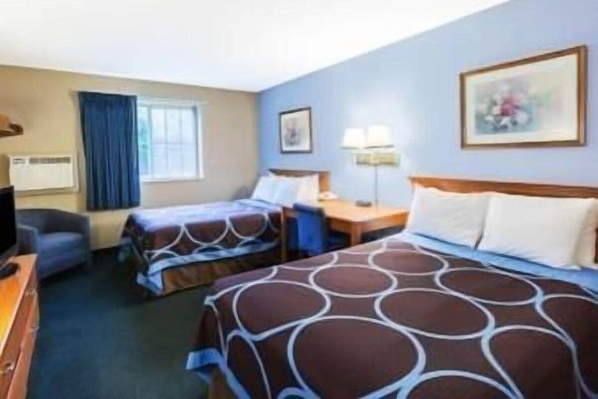 A hotel room with two beds, a desk, a chair, a window with curtains, a lamp, and a painting on the wall.