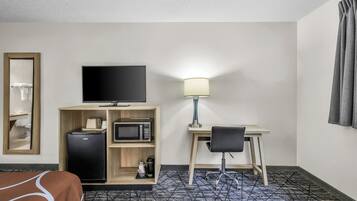 Habitación de hotel con cama, escritorio con silla, televisión, refrigerador, microondas y lámpara.