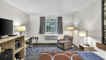 Habitación de hotel con cama, escritorio, silla, televisor, aire acondicionado y ventana con cortinas.